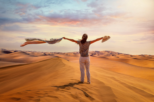 A tourist woman stands in the Red Desert of Dubai waving her scarf in the hot air, United Arab Emirates