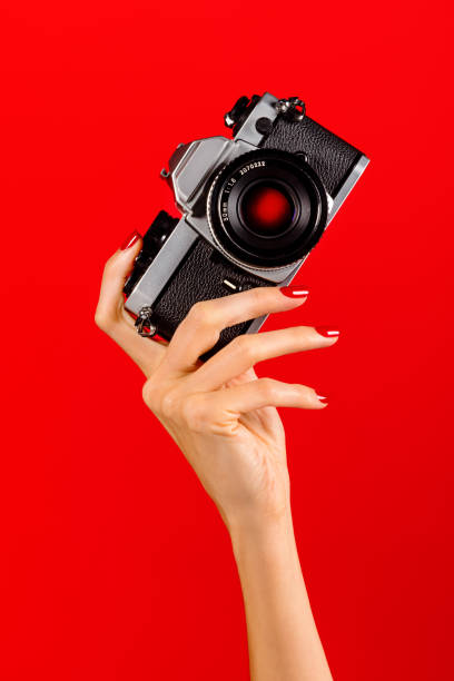 Anonymous slim female with manicured polished nail fingers holding vintage reflex camera with shutter eye in hand in light against red background