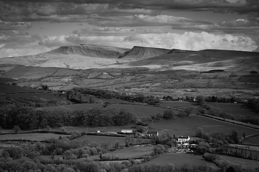 Dive into the timeless allure of the Brecon Beacons with this captivating black and white photograph taken from Tally Woods. Behold the majestic silhouette of the Black Mountains, their peaks reaching for the sky, accentuated by a dramatic canvas of swirling clouds. This striking composition captures the raw, untamed beauty of the natural landscape. Ideal for conveying a sense of awe and wonder, this image is a testament to the grandeur of the Brecon Beacons.