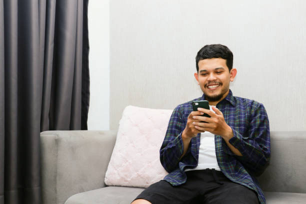 istock Happy young man sitting on a gray sofa and holding a mobile phone. Guy browsing internet, surfing web, using app, smiling expression 1774548299