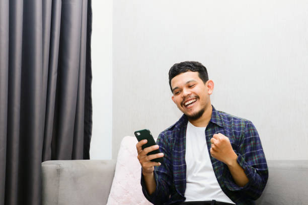 istock Happy young man sitting on a gray sofa and holding a mobile phone. Guy browsing internet, surfing web, using app, smiling expression 1774548321