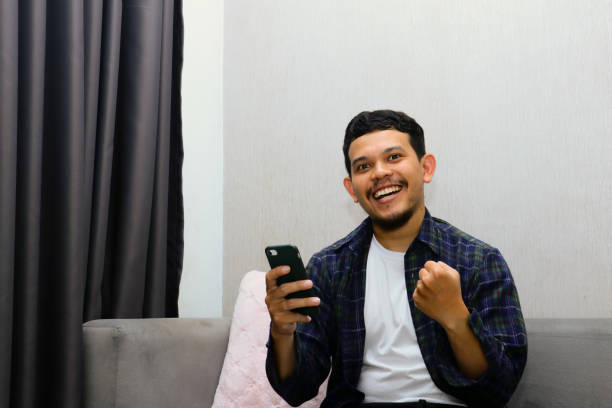 istock Happy young man sitting on a gray sofa and holding a mobile phone. Guy browsing internet, surfing web, using app, smiling expression 1774548343