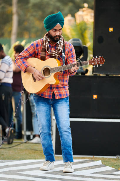 молодой индийский сикх в бирюзовом головном уборе, красной рубашке и синих джинсах играет на гитаре на открытом воздухе - new delhi audio стоковые фото и изображения