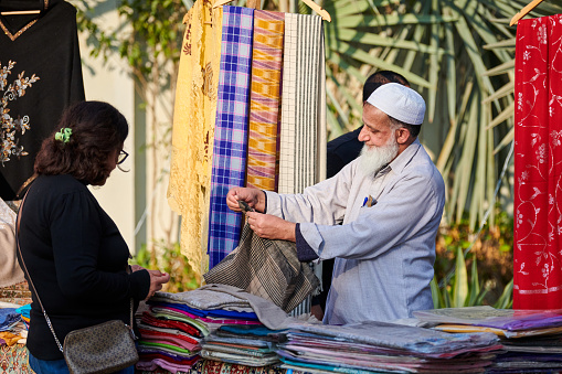 New Delhi, India - 10.12.2022 - Outdoor public park. Old seller of textile and carpet shows goods to woman at outdoor fair, bearded muslim salesman in taqiyah cap shows examples of fabrics for sale on indian market