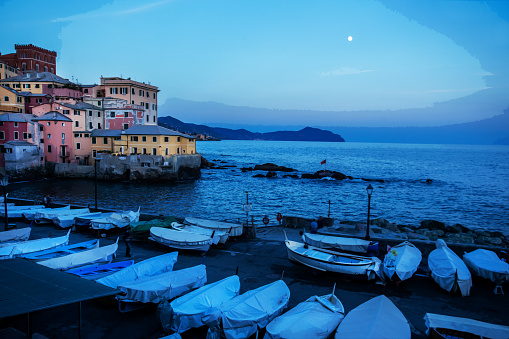 Fantastic sunset View from Boccadasse old  fishing  village in Genoa suburb   andf moon in sky , many boats sheltering on ground\nPink and yellow fisher's houses on cliff. Blurred motion people.