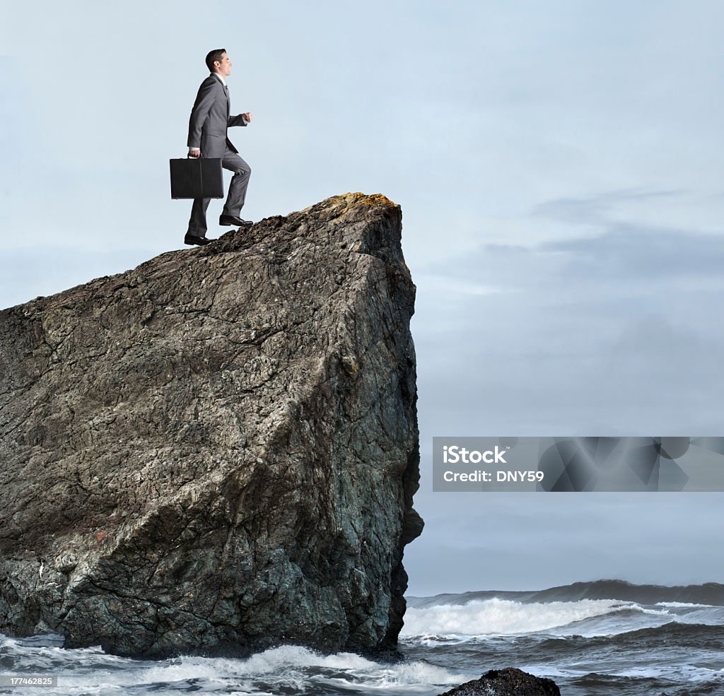 Businessman Climbing A Cliff Overlooking The Ocean A businessman climbing a cliff overlooking the ocean. He is holding a briefcase.  Small waves are breaking against cliff below. There is room for text. Adult Stock Photo