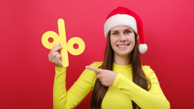 Merry pretty young woman wear cozy yellow sweater and Santa Claus hat holding big paper yellow percent mark, posing isolated over pastel red studio background wall. Happy New Year 2024 holiday concept