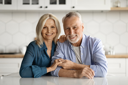 Portrait of happy loving senior couple in casual outfits posing together at cozy kitchen interior, mature spouses sitting at table, embracing and smiling at camera, enjoying time at home, copy space