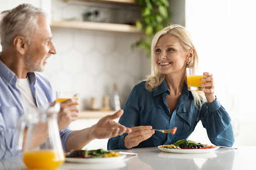 Breakfast Together. Happy Senior Spouses Eating Meal And Drinking Orange Juice In Kitchen, Married Elderly Couple Chatting And Laughing While Sitting At Table Enjoying Spending Time At Home, Closeup