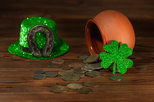 St Patricks day concept with pot with scattered old coins, green hat, horseshoe and shamrock on vintage wooden background, close up.