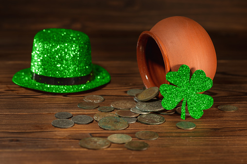 St Patricks day concept with pot with scattered old coins, green hat and shamrock on vintage wooden background, close up.