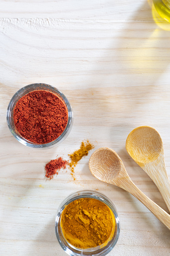 Spain. Top-down view of little glass containers with turmeric and paprika next to wooden spoons on a wooden table. Paprika is a red or orange-colored powdered spice obtained from the drying and grinding of red peppers. Turmeric is a bright yellow spice derived from the root of the Curcuma longa plant.