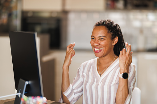 African female freelancer anticipating with fingers crossed while working on computer from her home office.