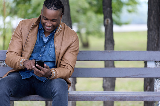 Portrait of a confident young African American student in stylish clothes, sitting on a bench in a city park with a smartphone. Black student reads a fun e-book in the park with interest