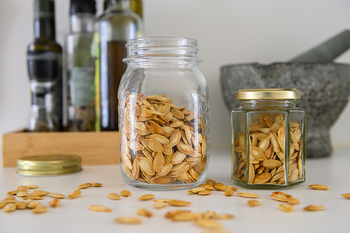 Two glass jars with roasted pumpkin seeds on a kitchen table. Oil and vinegar bottles and a mortar and pestle in the background
