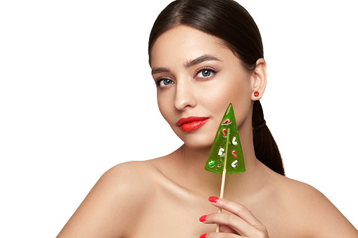 Beauty model girl eating Christmas tree shape lollipop.  Beautiful young woman with clean fresh skin. Girl beauty face care. Facial treatment. Cosmetology, beauty and spa. Red nails manicure