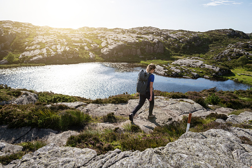 TOURIST MAN ON TOP OF A ROCK WITH BACKPACK AND HAT LOOKING WITH BINOCULARS IN A BEAUTIFUL LANDSCAPE WITH MOUNTAINS NEXT TO A LAKE, RIVER OR SEA. TOURISTIC VIEWPOINT. TRAVELER MAN CONCEPT.