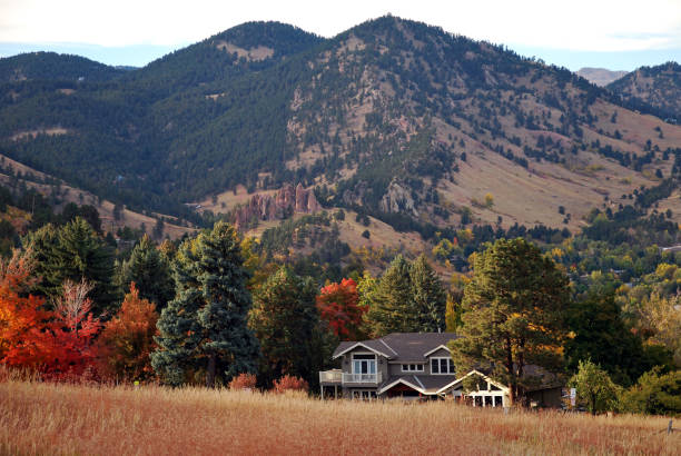 As the sun hovers just above the horizon, nature's canvas comes alive in a symphony of hues. Majestic peaks embrace a quaint prairie house, surrounded by vibrant autumnal trees and rustic megalithic stones. A golden meadow stretches gracefully, whispering tales of time amidst tranquil twilight in the Rockies.