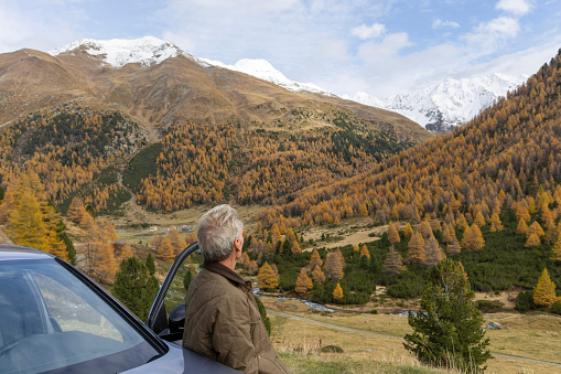 He leans against car and looks across a valley to larch trees on a mountain slope in autumn