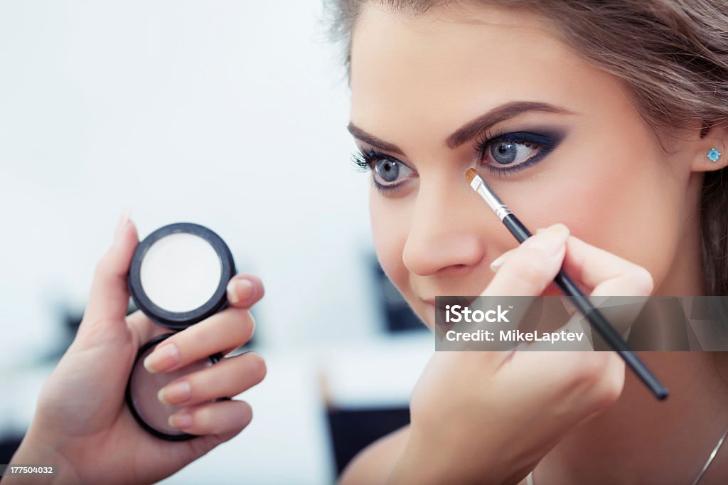 A makeup artist putting eyeshadow on a woman "Make-up artist applying white eyeshadow in the corner of model's eye and holding a case with eyeshadow on background, close up" Adolescence Stock Photo