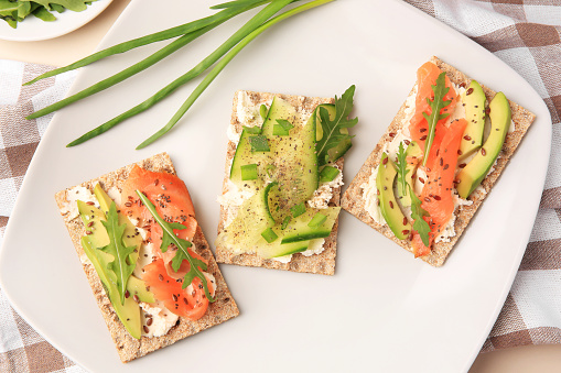 Fresh crunchy crispbreads with cream cheese, cucumber, green onion, salmon and arugula on plate, flat lay