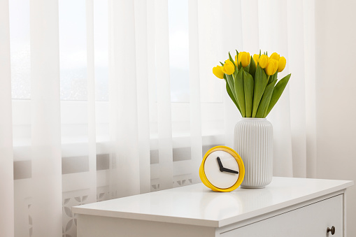 Spring interior. Beautiful yellow tulips and clock on white chest of drawers near window, space for text