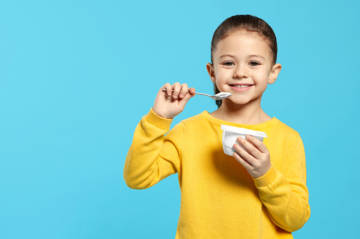 Girl eating tasty yogurt on light blue background, space for text