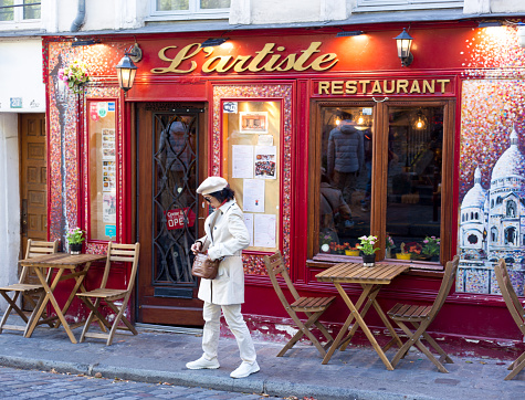 Paris, France: A woman dressed all in white (including a white beret) stands outside the bright red Montmartre restaurant “L’Artiste.”