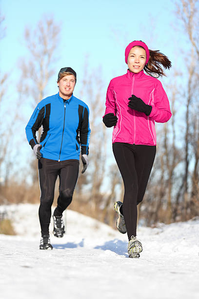 Young couple running dressed warmly in fleeces and gloves jogging in sunshine across winter snow in the countryside.
