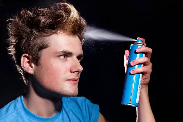 "Young man sprays his hair with aerosol hairspray, droplets visible in the spray at close up."