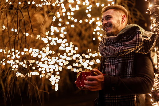 Portrait of young man standing by the glistening Christmas lights at the park, holding a Christmas gift box and waiting for someone to meet.