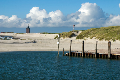 View of the West Beach of Wangerooge Island with Old Westtower (Alter Westturm) and New Lighthouse (Neuer Leuchtturm). Sand dunes with beachgrass and the jetty in the harbor between deep blue sea and a cloudy blue sky. Taken in Wangerooge Island, Ostfriesland, Lower Saxony, Germany, Europe.
