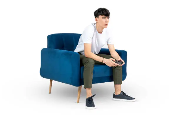 Teenager playing videogames, with a surprised expression, sitting on a blue armchair, isolated on white background. Clipping Path in the file.