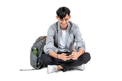 Student texting, using his smartphone sitting on the floor, isolated on white background.