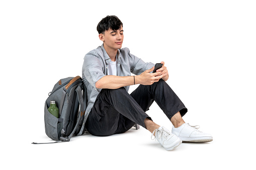 Student texting, using his smartphone sitting on the floor, isolated on white background.