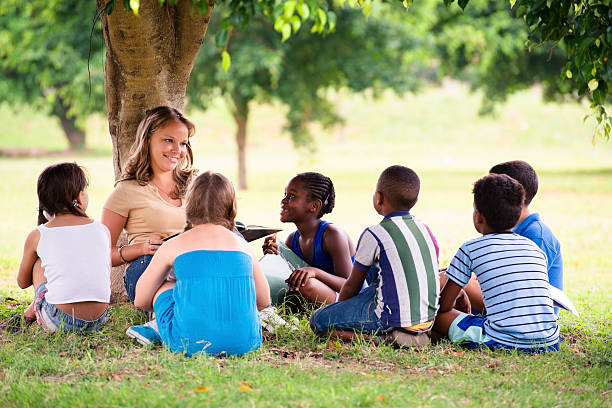 Sitting female teacher surrounded by school-aged children Children and education, young woman at work as educator reading book to boys and girls in park travel9 stock pictures, royalty-free photos & images