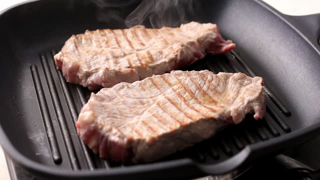 Pieces of raw meat are turned over on a grill pan using tongs.