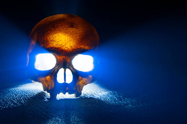 A scary glowing skull sits on stone surface and emits fog and light. The bone glows a warm yellow against a cool blue background. Rough textures can been seen on both the skull and the stone on which it sits.