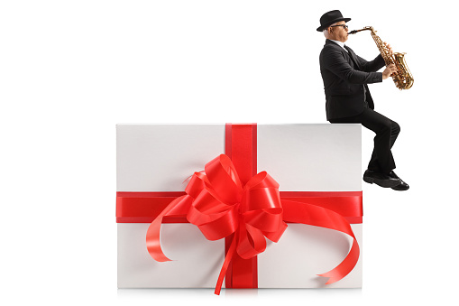 Full length profile shot of a male artist playing a sax and sitting on a present isolated on white background