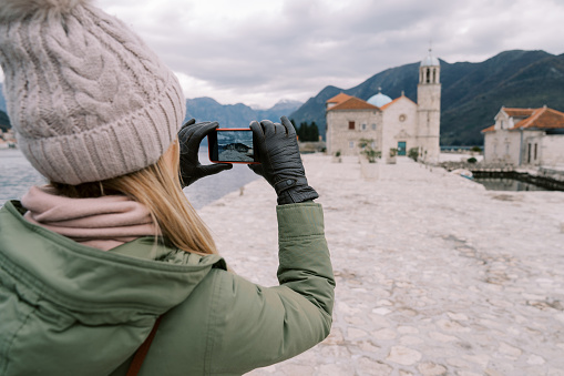 Young woman takes a picture of the Church of Our Lady on the Rocks with her smartphone. Back view. Montenegro. High quality photo
