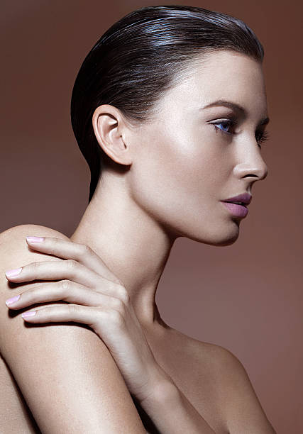 Portrait of beautiful young woman in profile "Portrait of young stylish woman with the collected wet hair, tanned skin on the brown background. In profile, with a hand on his shoulder, naked shoulders and neck. Natural make-up, blue eyes, beautiful, clear skin, light pink lipstick and nail polish." slicked back hair stock pictures, royalty-free photos & images