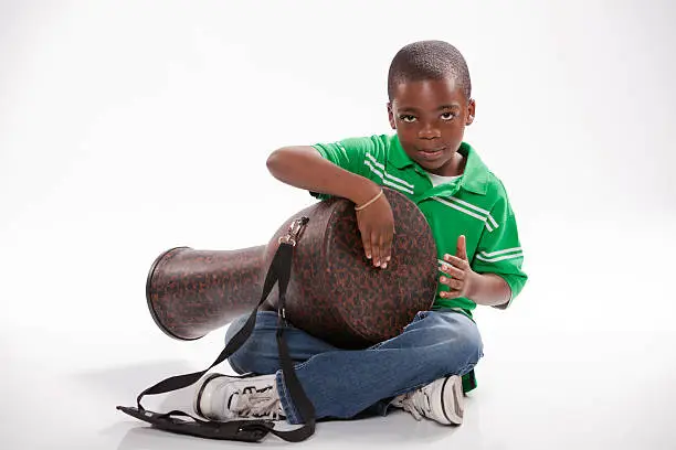 A small isolated African American male child in a green shirt studying how to play a djembe drum against a white background.
