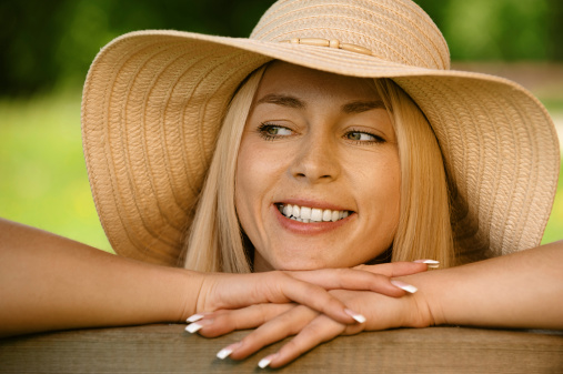 Pretty smiling young woman in hat sitting on wooden bench close up, against green of summer park.