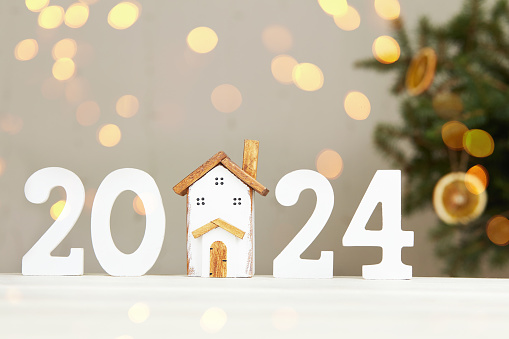 Happy New Year 2024 and Merry Christmas greeting card. Toy wooden house, white numbers 2024, Christmas tree, bokeh lights.