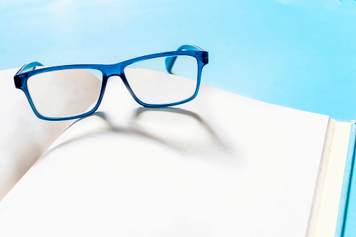 Blue glasses on the book. Eyeglasses and book on blue background. Glasses and book place on table in the living room. There is space to put text.