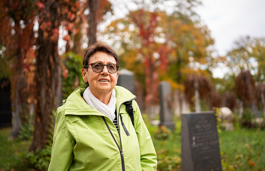 portrait of one senior adult woman walking through old cemetery in vienna on bright day in autumn, colorful trees in background