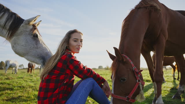 SLO MO Smiling Young Female Rancher in Red Plaid Shirt Sitting Near Horses Grazing on Meadow During Summer