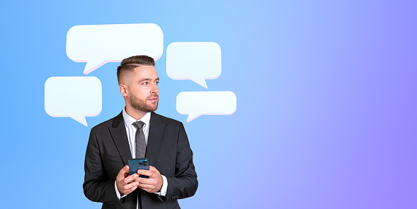 Portrait of handsome young European businessman in elegant suit with smartphone looking sideways at copy space standing over blue background with speech bubbles. Concept of social media