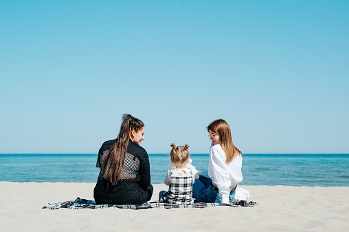 Back view of a woman, girlfriend, and little girl, daughter, spending time together on beach by sea, sitting on a blanket.