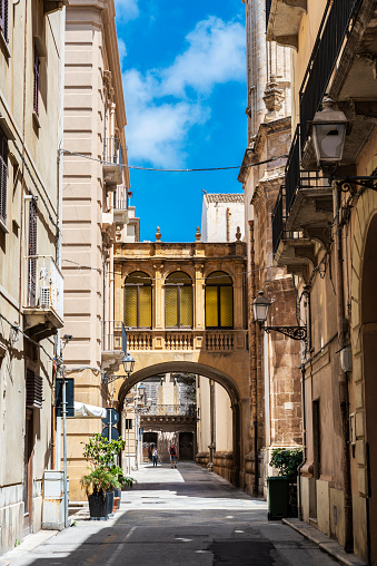 Street of classic buildings with an elevated exterior walkway connecting separate buildings and people around in the old town of Marsala, Sicily, Italy